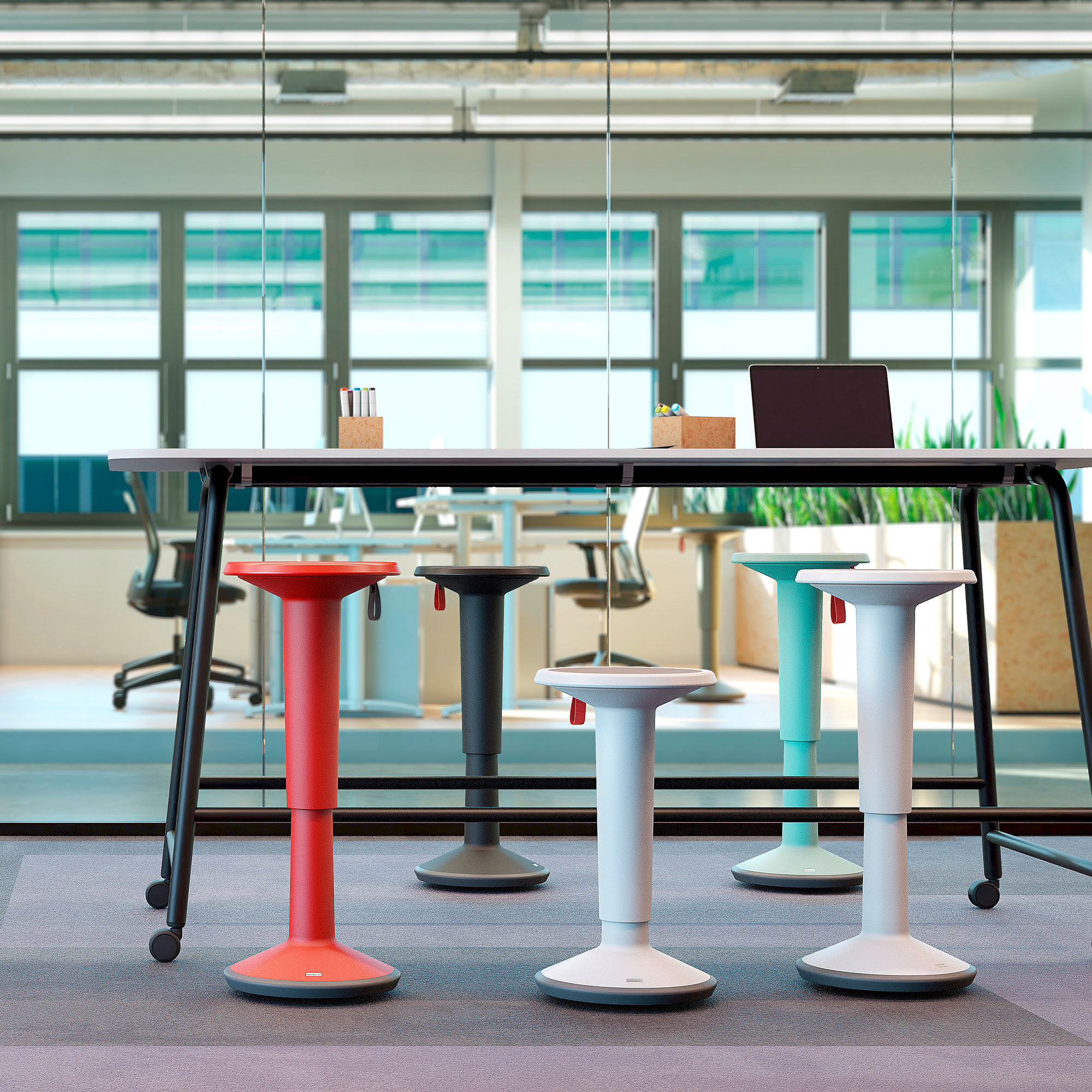 Ein moderner Büroraum mit einem langen Tisch und vier bunten Stehhilfen in Rot, Schwarz, Weiß und Blau. Im Hintergrund sind Bürostühle und Pflanzen zu sehen.