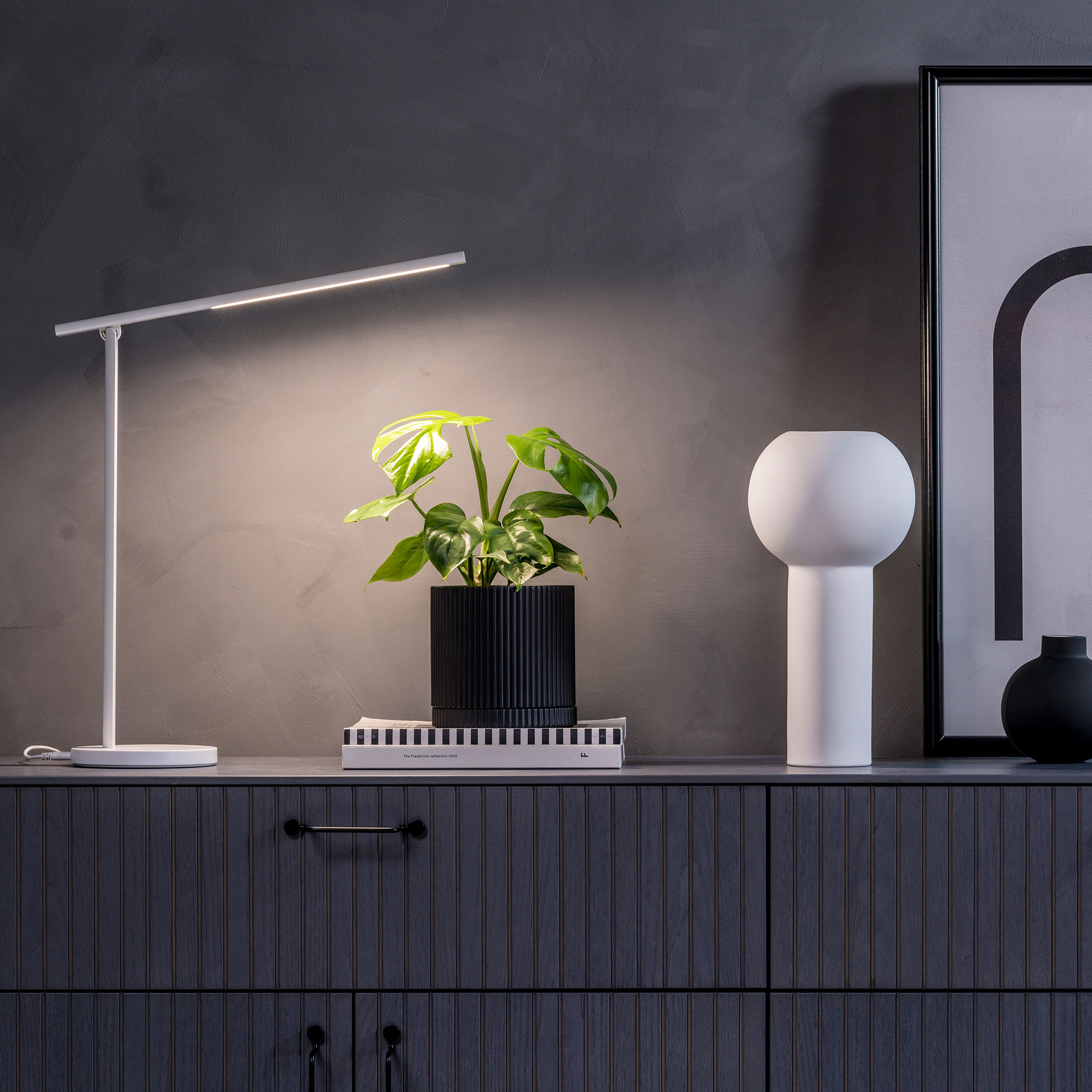 Eine moderne Tischlampe, eine Pflanze in einem schwarzen Topf, ein weißer dekorativer Behälter und ein gerahmtes Bild auf einer Kommode vor einer grauen Wand.