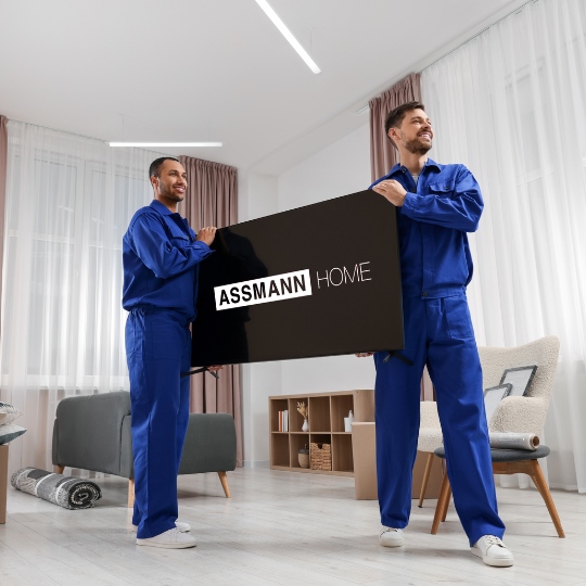 Zwei Monteure in blauer Arbeitskleidung tragen großen schwarzen Fernseher mit ASSMANN HOME Aufschrift in moderne Wohnung