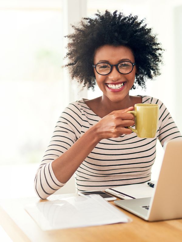 Lächelnde Frau mit Brille und gestreiftem Shirt trinkt Kaffee am Schreibtisch mit Laptop.