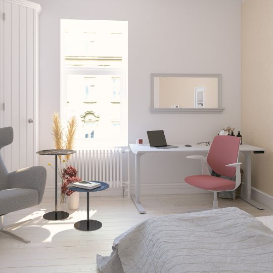 Ein helles Zimmer mit einem Schreibtisch, einem rosa Stuhl, einem Spiegel, zwei Tischen und einer gemütlichen Ecke mit einem grauen Sessel.