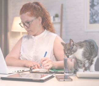 Eine Frau mit roten Haaren sitzt am Schreibtisch und arbeitet am Laptop. Neben ihr steht ein Glas Wasser, und eine Katze schnuppert daran.