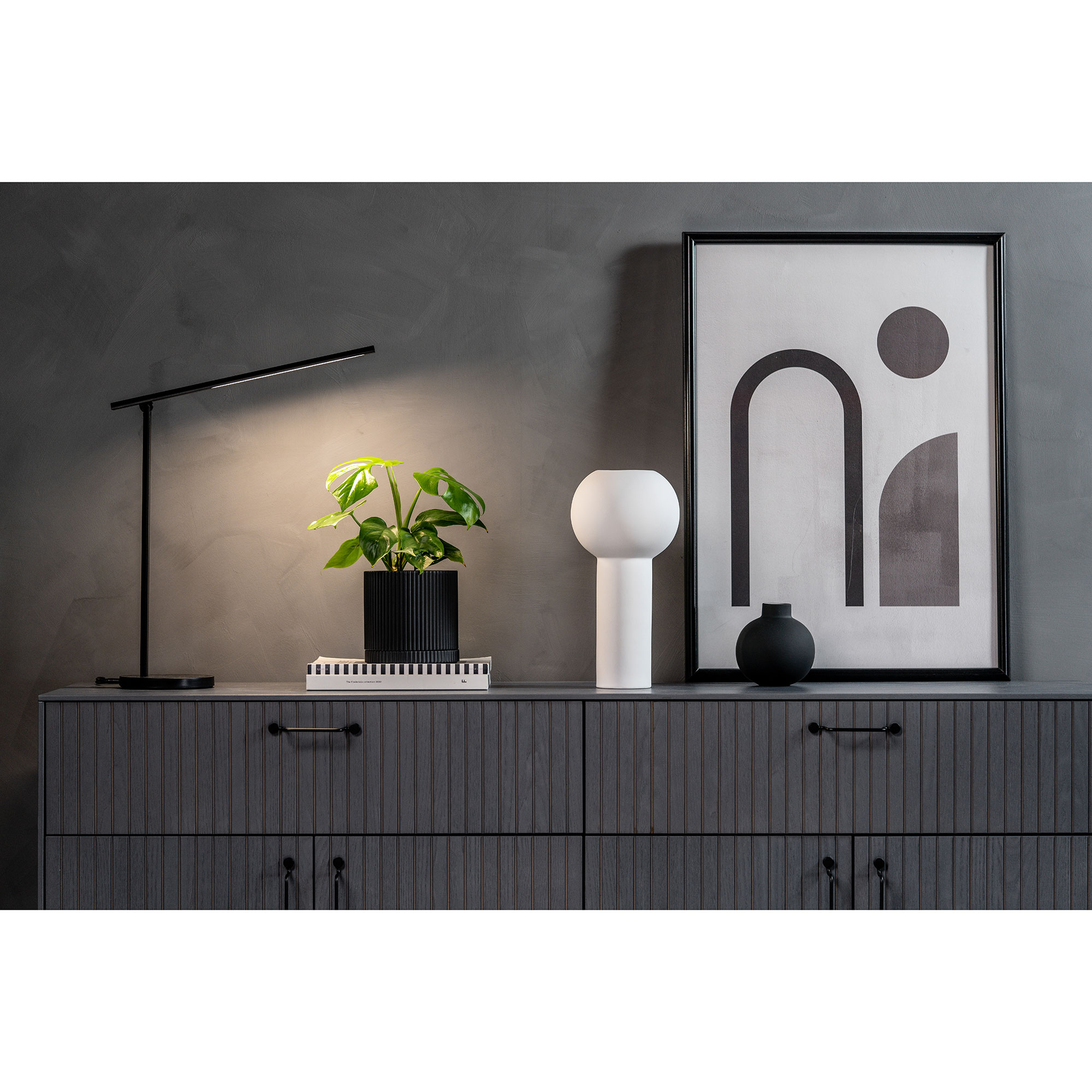 Ein moderner Raum mit einer Lampe, einer Pflanze in einem schwarzen Topf, einem weißen Leuchten und einem abstrakten Grafikdruck an einer grauen Wand.