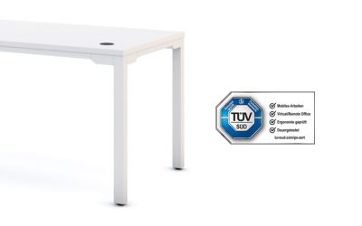 Ein weißer höhenverstellbarer Schreibtisch, das große Modell ASSMANN Piacetta mit vier Tischbeinen. Neben dem Tisch ist das TÜV SÜD Zertifikat zu sehen.