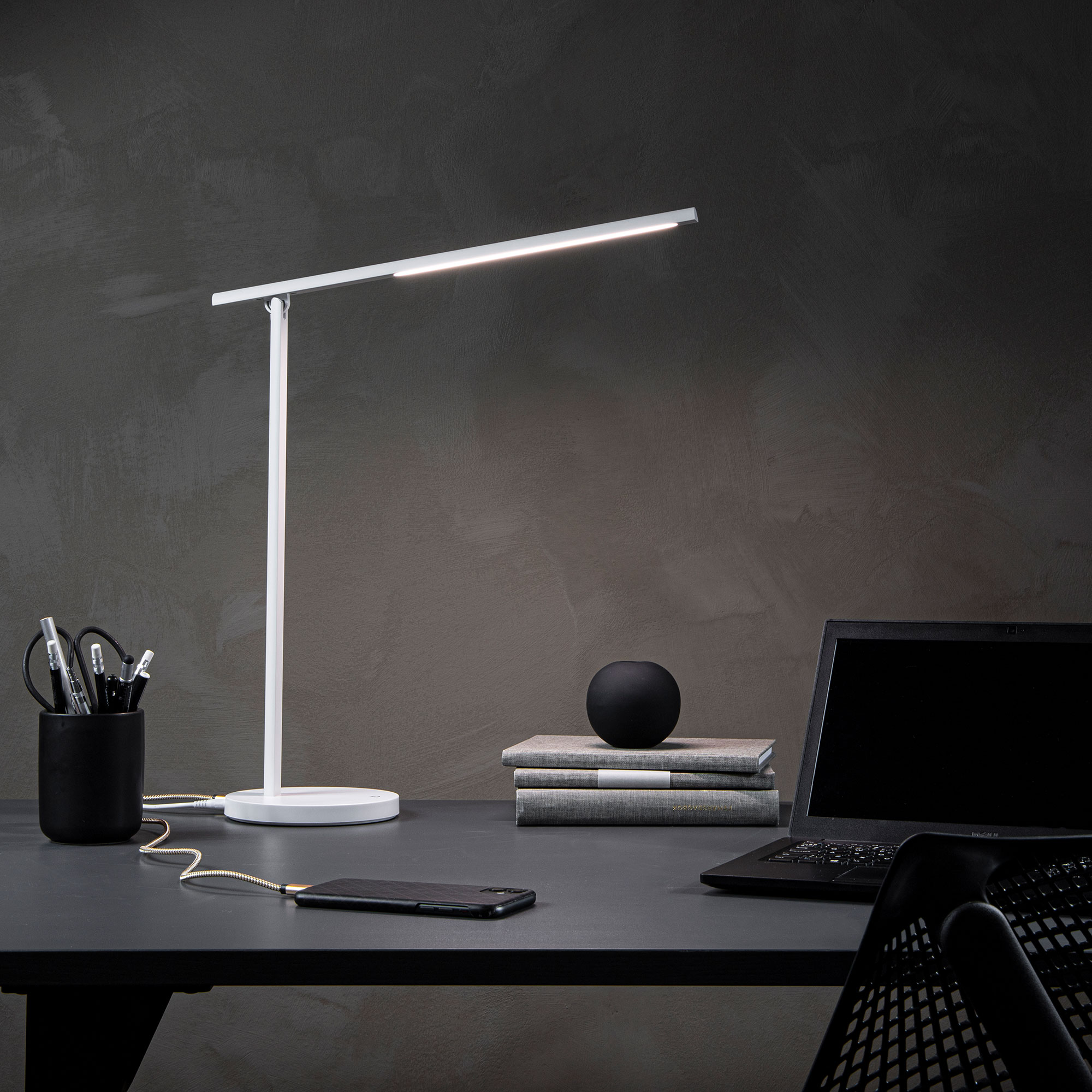 Ein moderner Schreibtisch mit einer weißen Tischlampe, einem schwarzen Stiftehalter, einem Smartphone, mehreren Notizbüchern und einem Laptop.