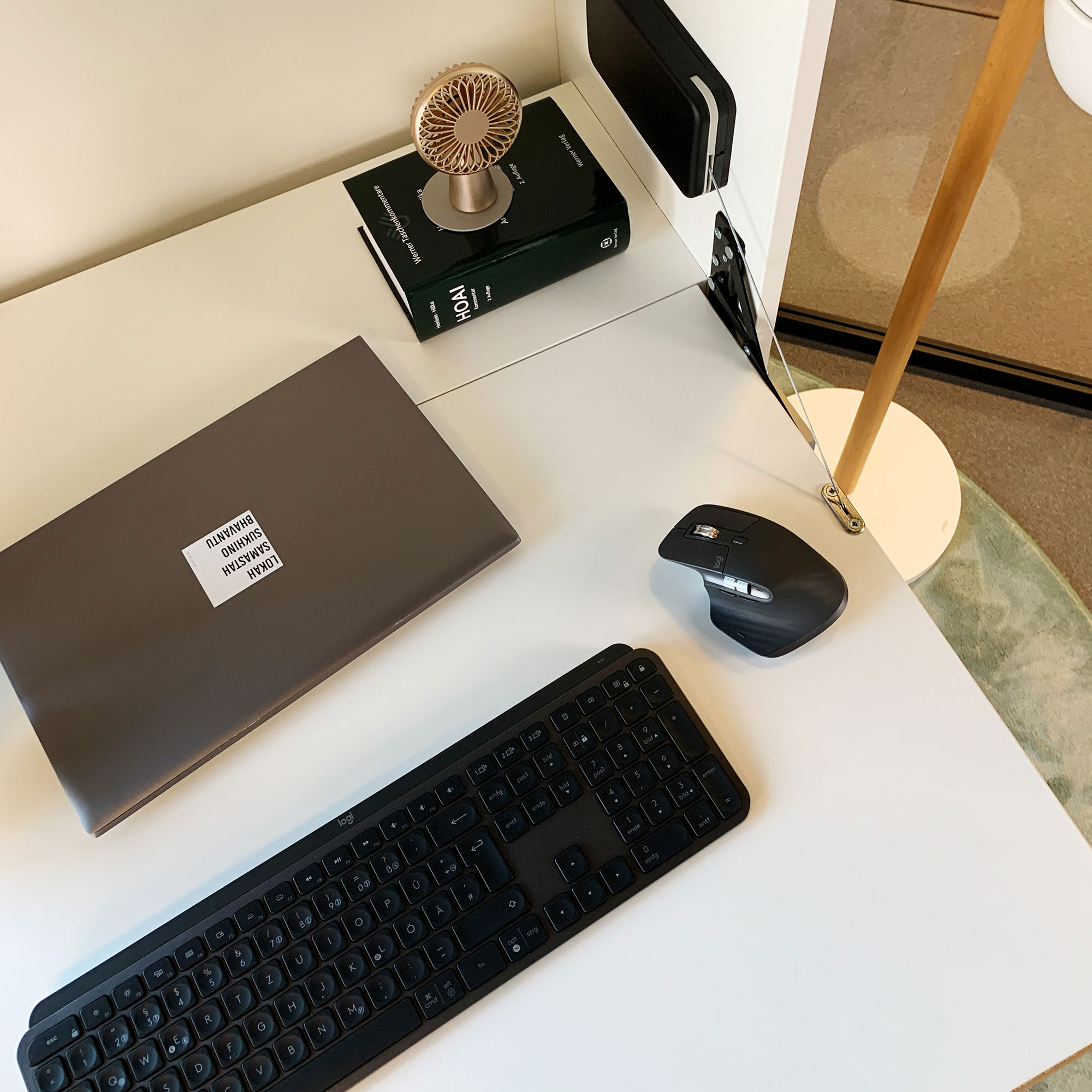 Ein Schreibtisch mit einem Laptop, einer Tastatur, einer Maus, einem kleinen Ventilator und einem Buch.