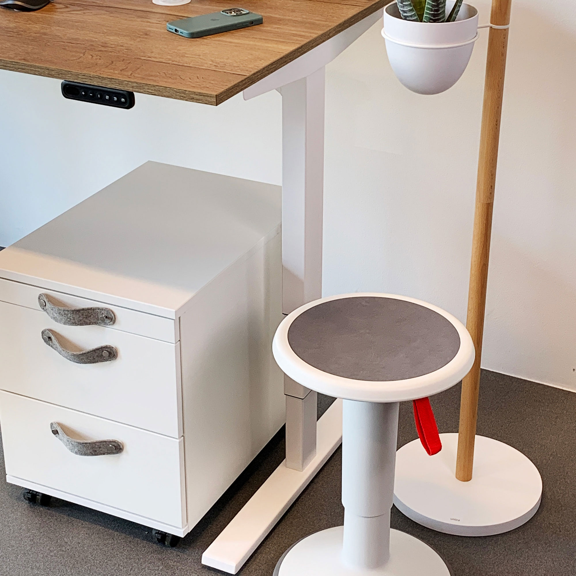 Ein Schreibtisch mit Holzoberfläche, einem weißen Rollcontainer, einem Hocker mit grauer Sitzfläche und einer Pflanze in einem weißen Topf.