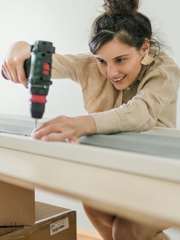 Eine Frau verwendet eine Akku-Bohrmaschine, während sie an einem Tisch arbeitet. Im Hintergrund sind Kartons zu sehen.