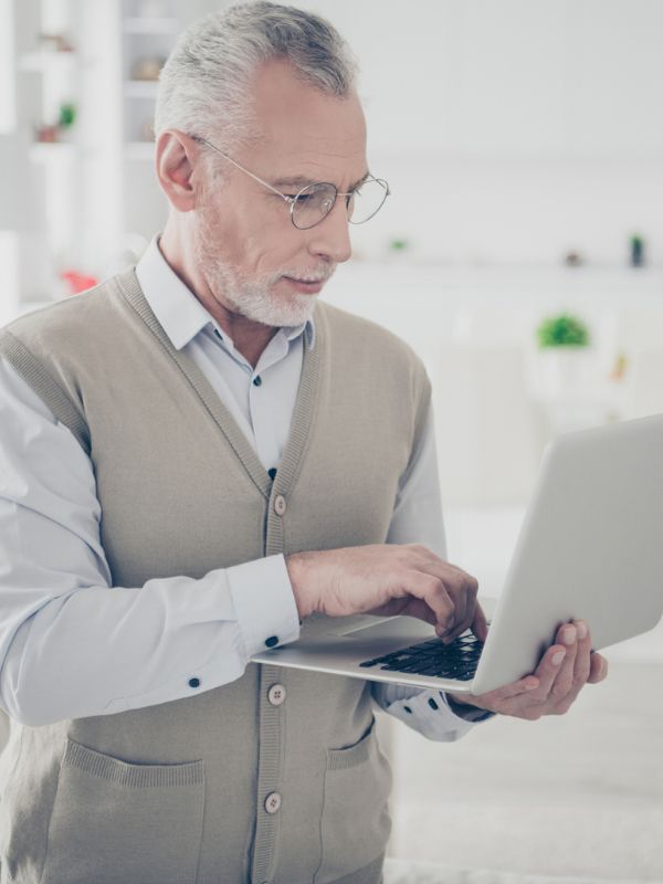 Ein älterer Mann mit Brille in einem grauen Pullover steht und tippt auf einem Laptop.